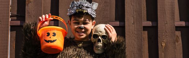 カメラで微笑みながらハロウィーンのバケツと頭蓋骨を持つ狼の衣装を着たアジアの少年,バナー - 写真・画像