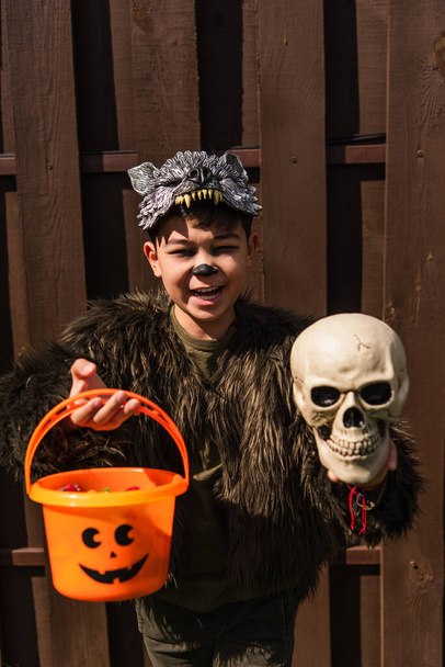 boldog ázsiai fiú vérfarkas jelmezben kezében kísérteties koponya és halloween vödör, miközben a kamera - Fotó, kép