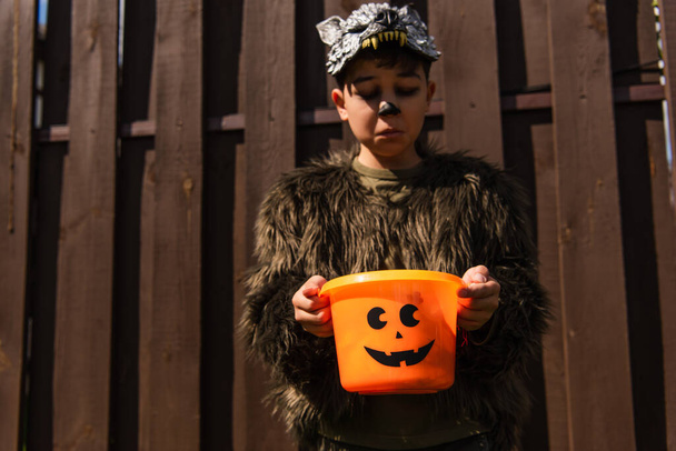 uražený asijský chlapec v vlkodlačí kostým při pohledu do prázdného halloween vědra - Fotografie, Obrázek