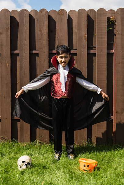 full length of asian boy in vampire costume holding cloak near skull and halloween bucket on grass - Fotó, kép