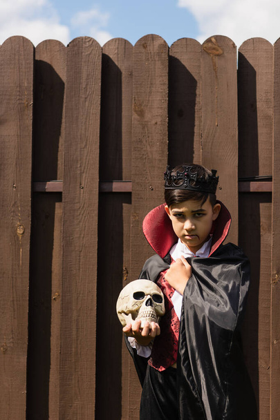 serious asian boy in vampire costume wrapping in cape while standing with skull near wooden fence - Photo, Image