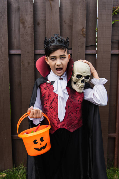 ázsiai fiú halloween jelmez mutatja dühös grimasz és sikoltozik a koponya és trükk vagy kezelésére vödör - Fotó, kép