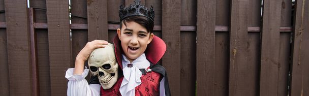 feliz asiático chico en vampiro traje mirando cámara mientras sostiene cráneo, bandera - Foto, Imagen