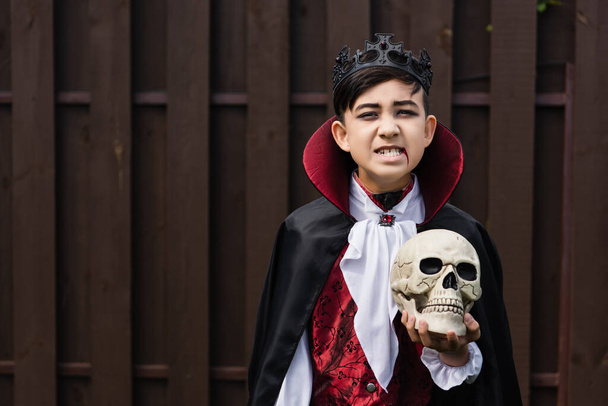asiático chico en vampiro rey traje gruñendo mientras celebración cráneo y mirando cámara - Foto, Imagen