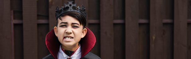 Vampir kral kostümü giymiş Asyalı çocuk korkunç bir somurtma ve pankart gösterirken hırlıyor. - Fotoğraf, Görsel