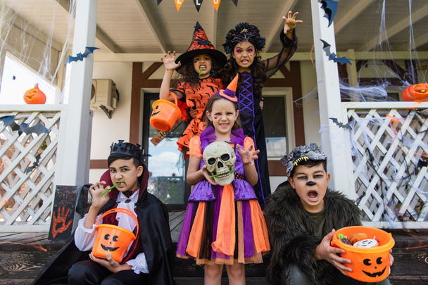 multiethnic friends in halloween costumes holding buckets of candies and grimacing near decorated cottage - Photo, Image
