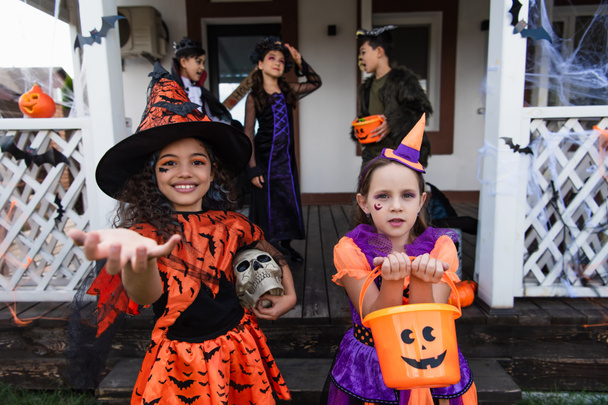 smiling girl in witch costume standing with outstretched hand near friend with halloween bucket  - Фото, зображення