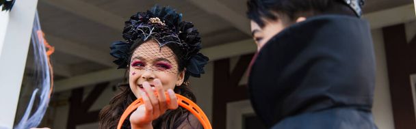 Lächelndes Mädchen im Halloween-Kostüm mit Blick auf verschwommene asiatische Freundin im Freien, Banner  - Foto, Bild