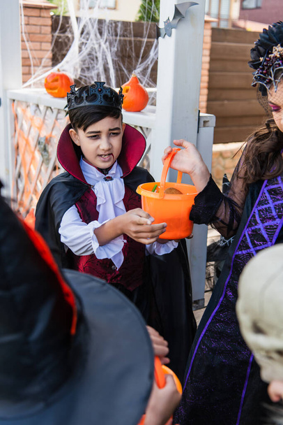 Asian boy in vampire costume holding bucket near friends during halloween celebration outdoors - Foto, afbeelding