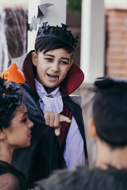 aziatische jongen in halloween vampier kostuum wijzend met vinger naar wazig vrienden buiten - Foto, afbeelding