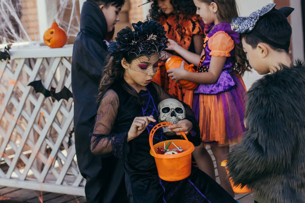 Shocked girl in costume holding bucket with candies near asian friend during halloween celebration outdoors  - Φωτογραφία, εικόνα