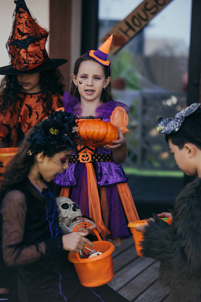 Preteen girl holding halloween pumpkin near multiethnic kids with buckets outdoors  - Foto, afbeelding
