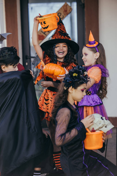 Cheerful girl in witch hat holding bucket near multiethnic friends during halloween celebration outdoors  - Foto, afbeelding