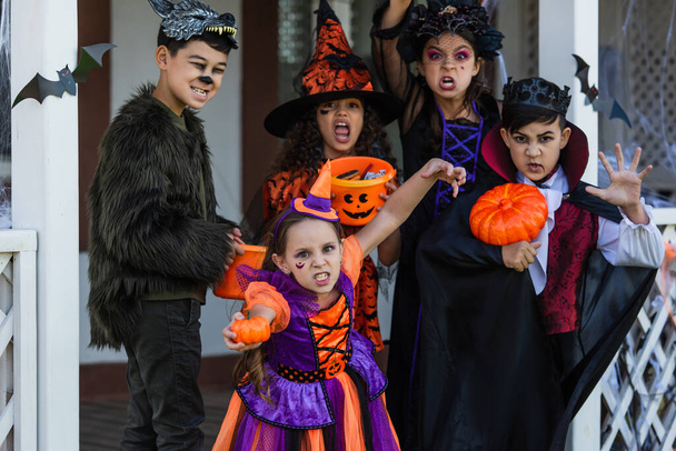 Multiethnic kids in halloween costumes grimacing at camera near house outdoors  - Photo, Image