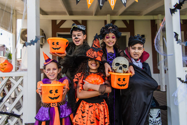 Happy multiethnic kids in halloween costumes holding buckets and pumpkin near house outdoors  - Photo, Image