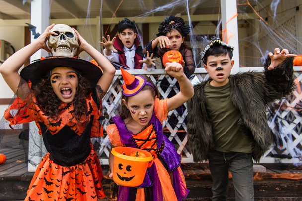 Preteen bambini multietnici con secchio e cranio smorzare fotocamera vicino amici e casa con arredamento di Halloween all'aperto  - Foto, immagini