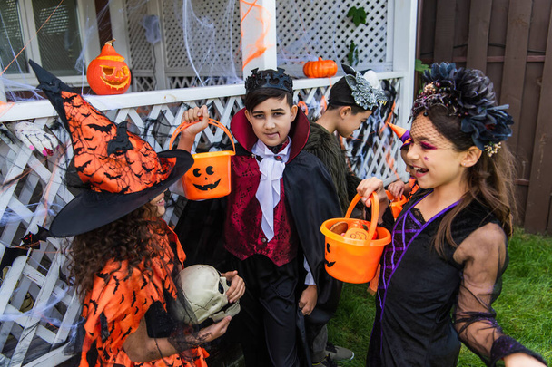Azji chłopiec w kostium patrząc na aparat w pobliżu wesołych przyjaciół z wiadro rozmowy podczas Halloween uroczystości na świeżym powietrzu  - Zdjęcie, obraz