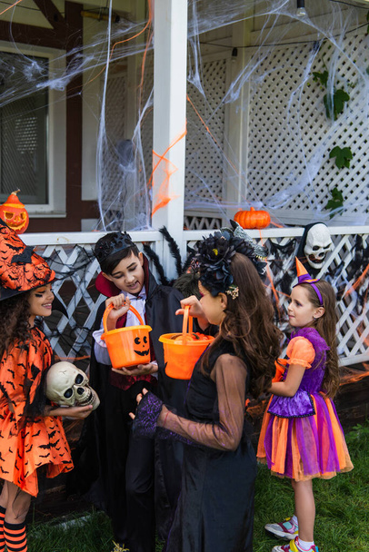Smiling preteen kids in halloween costumes looking at buckets with candies near interracial friends outdoors  - Фото, изображение