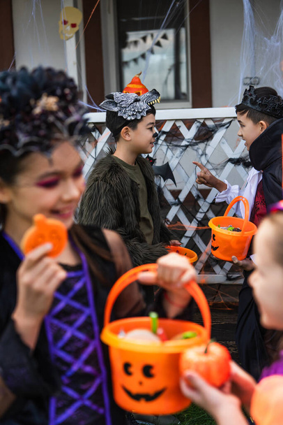 Aasialainen poika halloween puku pitäen ämpäri ja osoittaen sormella ystävää lähellä hämärtyneitä tyttöjä ulkona  - Valokuva, kuva