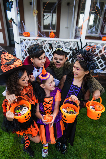 Cheerful multiethnic children in halloween costumes holding buckets with candies and pumpkin in backyard  - Photo, Image