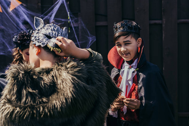uśmiechnięty azjatycki chłopiec w halloween kostium trzymając cukierki i patrząc na aparat w pobliżu przyjaciół w podwórku  - Zdjęcie, obraz