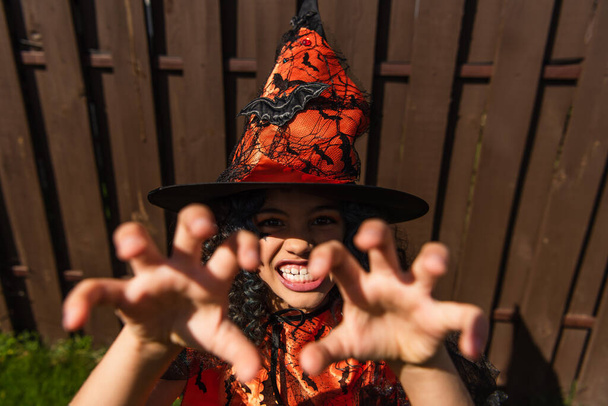 Vue grand angle de l'enfant en chapeau de sorcière halloween grimaçant à la caméra à l'extérieur  - Photo, image