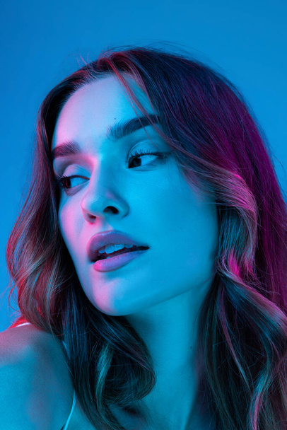 Portrait of young beautiful girl with curly long hair posing isolated over blue blackground in neon. Looks calm and relaxed. Health, beauty, fashion and ad concept. Closeup - Φωτογραφία, εικόνα
