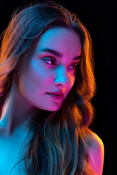Portrait of young beautiful girl with curly long hair posing isolated over dark blackground in neon. Looks happy and relaxed. Health, beauty, fashion and ad concept. Closeup - Valokuva, kuva