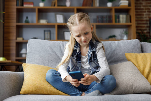 Portrait of cute little cute girl holding smartphone and resting at sofa at home. Choosing favorite music or cartoons,texting messages,browsing internet,watching video, playing games on mobile phone. - Fotoğraf, Görsel