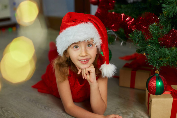 Happy Girl in Santa hat lying near christmas tree at home. Merry Christmas and Happy Holidays. - Fotoğraf, Görsel