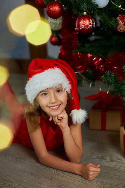Chica feliz en el sombrero de Santa que yace cerca del árbol de Navidad en casa. Feliz Navidad y Felices Fiestas. - Foto, imagen