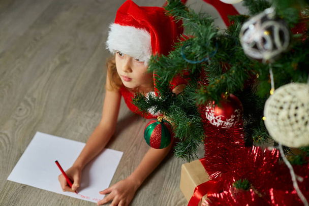 Meisje in Santa hoed schrijft brief naar de Kerstman in de buurt van kerstboom thuis. Vrolijk kerstfeest en fijne feestdagen. - Foto, afbeelding