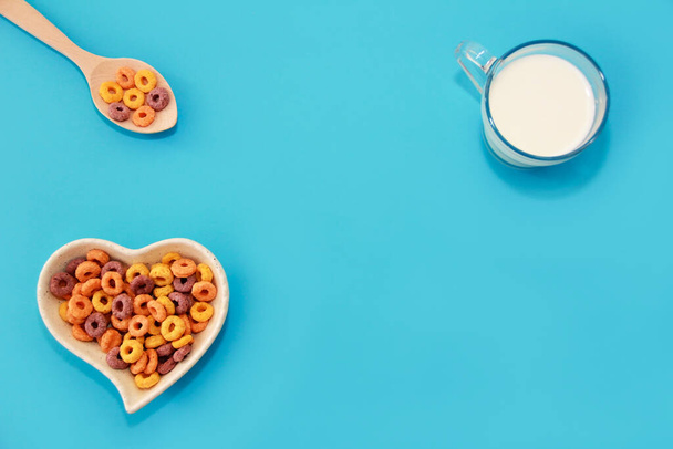 desayuno infantil seco anillos multicolores en un plato decorativo y una taza de vidrio con leche sobre un fondo azul - Foto, Imagen
