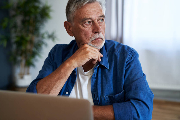 Senior homme caucasien assis à l'intérieur avec ordinateur portable et regardant loin - Photo, image
