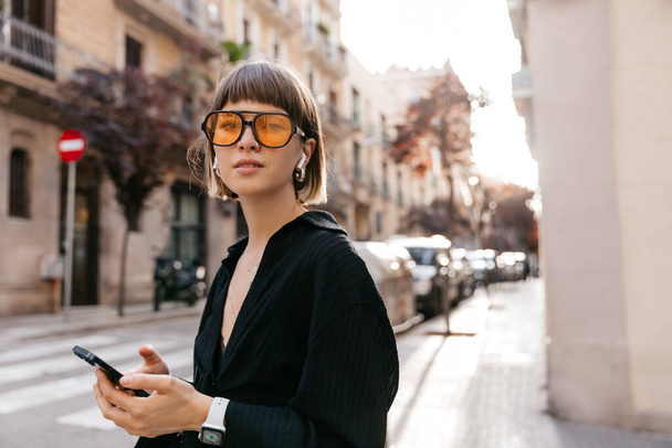 Spectacular charming lady with short hair wearing black shirt and stylish sunglasses listens music and using smartphone. Happy woman walking in the city in summer sunny evening  - Photo, Image