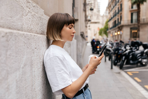 Profile photo of appealing stylish girl with short hair wears white t-shirt and jeans is using smartphone on city background. Attractive caucasian woman walking around the city with smartphone  - Foto, Imagem