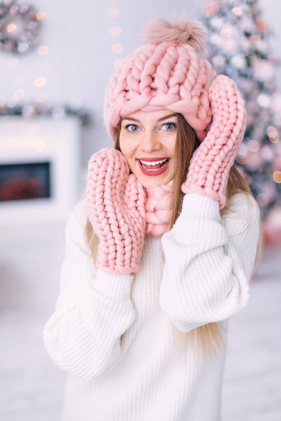 portrait of a girl in knitted winter clothes look down on a background of fireplace and Christmas tree. - Photo, Image
