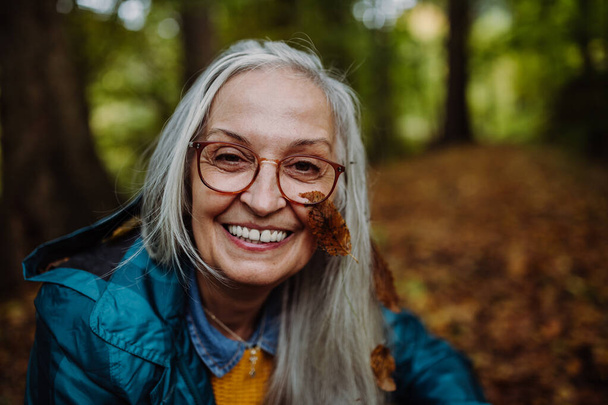Portrait of happy senior woman with a leaf on her cheek on walk outdoors in forest in autumn. - Foto, Imagen