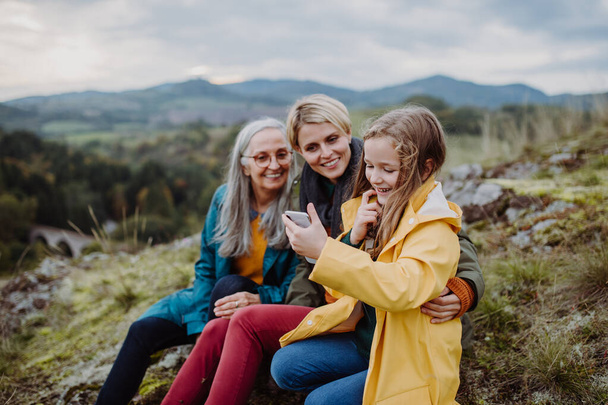 A small girl with mother and grandmother checking smartphone outoors on top of mountain. - Photo, Image