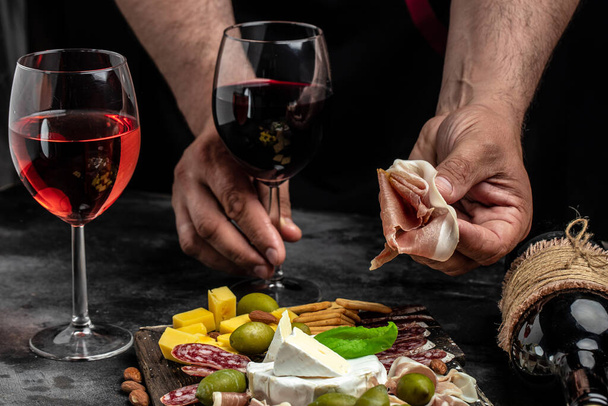 Kezek kezében egy pohár bor és egy fa tábla különböző típusú sajt és sonka, prosciutto, jamon szalámi, Antipasto Vacsora vagy aperitivo party. - Fotó, kép