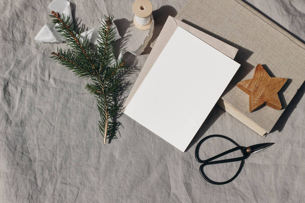 Christmas stationery. Blank greeting card, invitation mockup. Wooden star ornament, old books and spruce tree branches in sunlight. Beige linen tablecloth. Black vintage scissorsinter flat lay, top - Fotó, kép