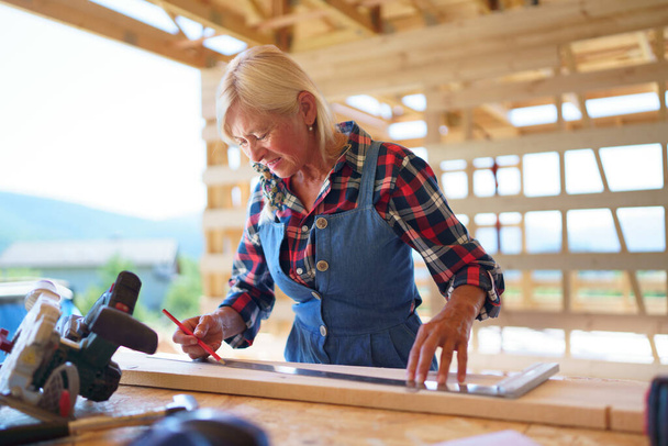 Senior woman working inside of her unfinished ecological renewable low energy sustainable wooden eco house. Concept of active independent women and seniors, eco house and healthy lifestyle. - Valokuva, kuva