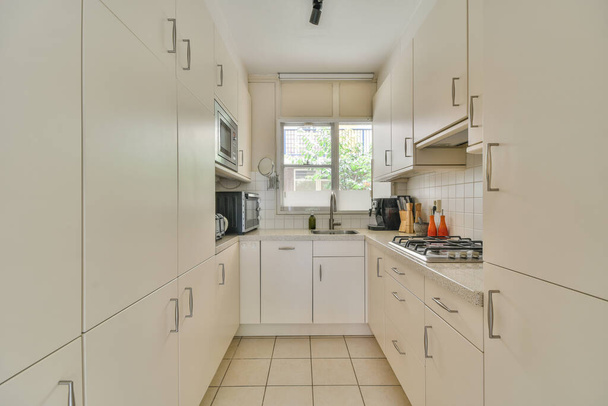Интерьер светлой кухни с современными шкафами и бытовой техникой расположен рядом со столовой в современной квартире в дневное время - Фото, изображение