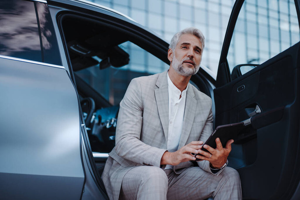 Businessman using tablet while charging a car at electric vehicle charging station, close-up. - Valokuva, kuva
