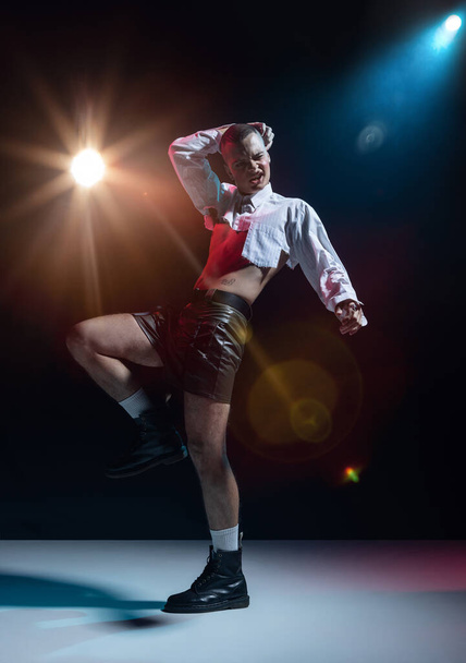 Portrait of stylish man in makeup posing in short white shirt and leather shorts on black background in neon. Expressing emotions. Concept of fashion, freedom of choice, lifestyle, lgbt, generation z - Photo, Image