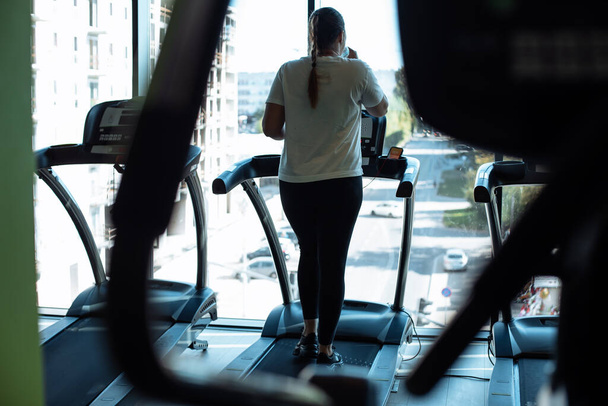 Plus size woman run on modern treadmill at fitness gym and look out window at transport traffic, back view. Overweight lady do cardio workout on exercise machine. Weight loss, sport lifestyle. - Fotó, kép