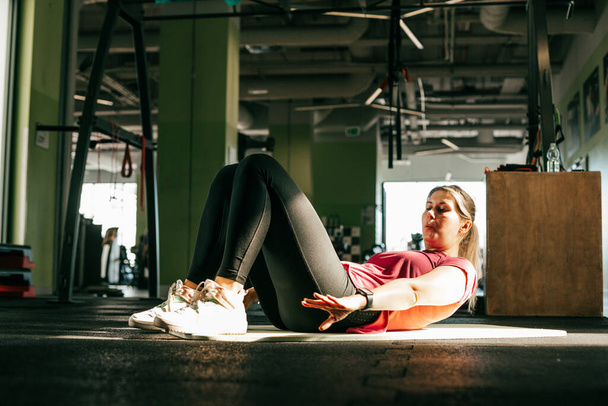 Overweight girl pump press in fitness gym lying on mat with bent knees and while exhaling reach arms to feet. Young plus size woman do cardio workout. Weight loss, abdominals, sport lifestyle. - Photo, Image