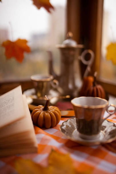 Herfst stilleven met kaarsen in de vorm van pompoenen, theekopje, herfstbladeren op het raam, theepot, gouden dienblad, vintage boek en glazen - Foto, afbeelding