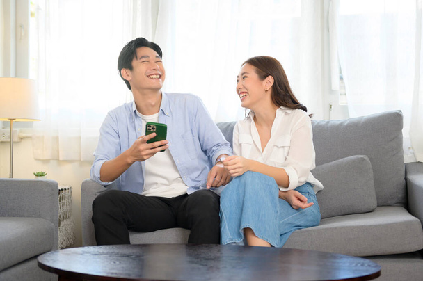 A Young smiling asian couple using smartphone in living room at home  - Fotografie, Obrázek