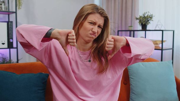Dislike. Upset girl showing thumbs down sign gesture, expressing discontent, disapproval, dissatisfied bad work at modern home apartment indoors. Displeased young woman in living room sitting on sofa - Foto, imagen